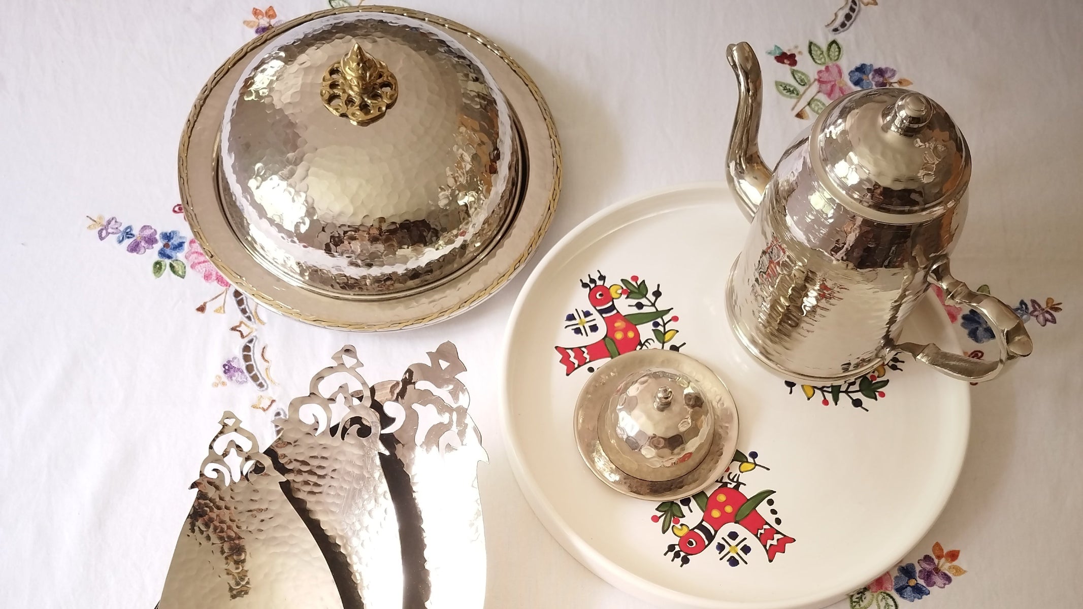 Image de couverture du blog. Cloche avec couvercle, théière, plateaux incurvés en cuivre martelé argenté et un plateau en céramique décor oiseau. Artisanat tunisien
